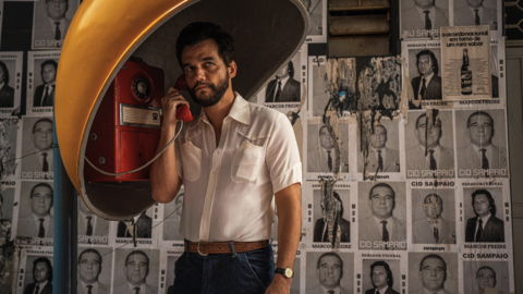 A man stands in a retro phone booth, holding a red receiver, against a wall of faded political posters.