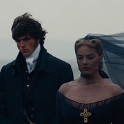 A man and woman stand solemnly on a foggy beach, the woman wearing a black dress and a flowing veil.