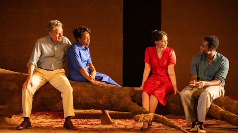 Four individuals are seated on a large branch in a warm, rustic setting, dressed in colorful clothing, engaged in conversation.