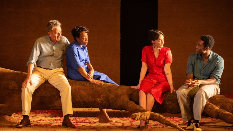 Four individuals are seated on a large branch in a warm, rustic setting, dressed in colorful clothing, engaged in conversation.