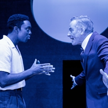 Two male actors engage in a heated discussion on stage, surrounded by deep blue lighting and a circular backdrop.
