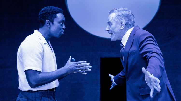 Two male actors engage in a heated discussion on stage, surrounded by deep blue lighting and a circular backdrop.