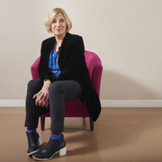 A person sits on a pink chair, wearing a black jacket and blue blouse, with black pants and stylish shoes, in a minimalist room.
