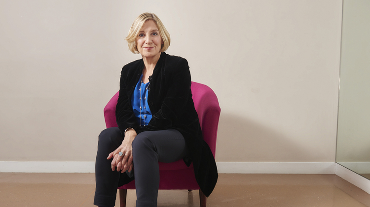 A person sits on a pink chair, wearing a black jacket and blue blouse, with black pants and stylish shoes, in a minimalist room.