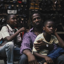 A group of children and a man sit in front of a stall filled with various types of shoes.