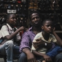 A group of children and a man sit in front of a stall filled with various types of shoes.