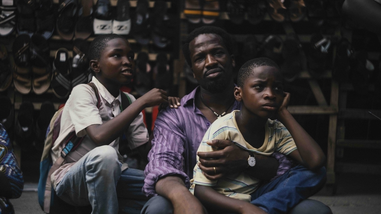 A group of children and a man sit in front of a stall filled with various types of shoes.