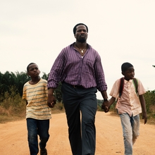 A person and two children walk hand in hand on a sandy path, surrounded by greenery under a cloudy sky.