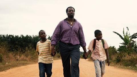 A person and two children walk hand in hand on a sandy path, surrounded by greenery under a cloudy sky.