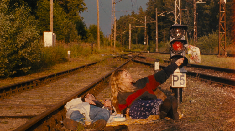 Two people relax on a picnic blanket by train tracks, with one holding a flag, surrounded by greenery.