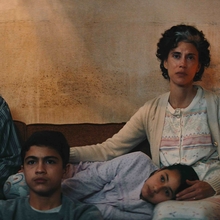A family of four sits closely together on a couch in a dimly lit room, surrounded by a rough-textured wall.