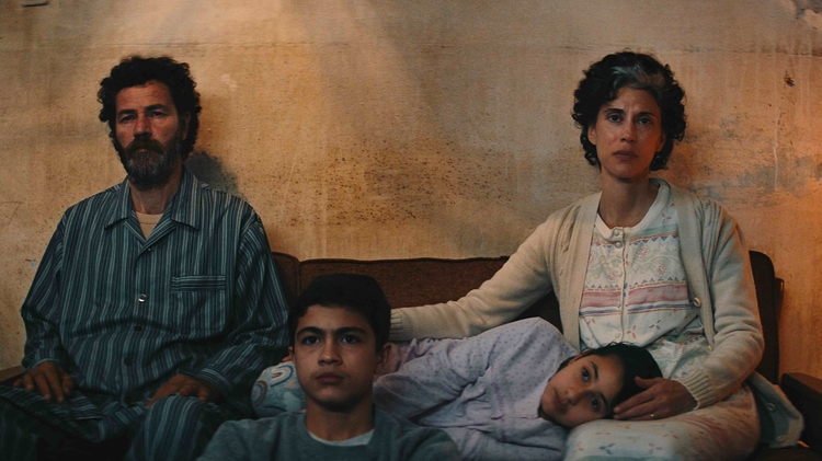 A family of four sits closely together on a couch in a dimly lit room, surrounded by a rough-textured wall.