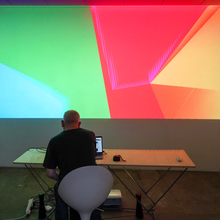 An image of a man, with his back turned to the camera, sat at a desk, manipulating an unseen controller for multi coloured lights and shapes projected onto the wall in front of him. The shapes are a warm mix of green, red, orange and yellow.