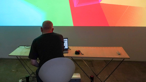 An image of a man, with his back turned to the camera, sat at a desk, manipulating an unseen controller for multi coloured lights and shapes projected onto the wall in front of him. The shapes are a warm mix of green, red, orange and yellow.