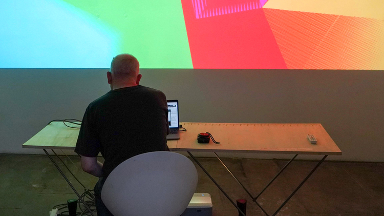 An image of a man, with his back turned to the camera, sat at a desk, manipulating an unseen controller for multi coloured lights and shapes projected onto the wall in front of him. The shapes are a warm mix of green, red, orange and yellow.