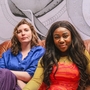An image of two women, Jess Bunyan and Euellla Jackson - they are sat together on a brown sofa, with an abstract pattern painted on the wall behind them. Jess wears blue, and Euella wears red. 