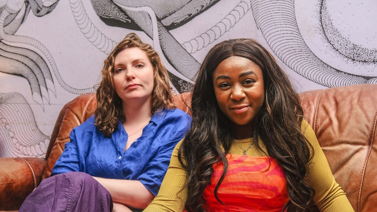 An image of two women, Jess Bunyan and Euellla Jackson - they are sat together on a brown sofa, with an abstract pattern painted on the wall behind them. Jess wears blue, and Euella wears red. 