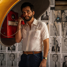 A person stands with a red rotary phone under a yellow hood, surrounded by faded political posters on a wall.