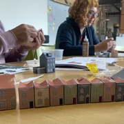 Two individuals are crafting paper models of colorful townhouses on a wooden table. A row of completed models lines the table, evoking a sense of creativity.