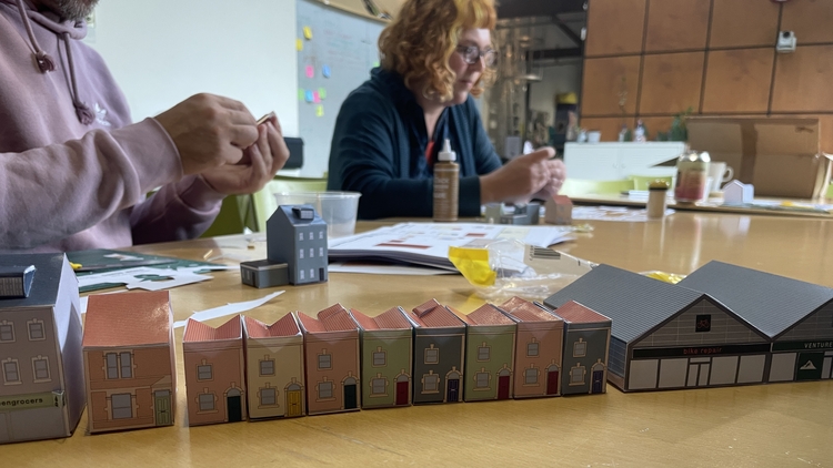 Two individuals are crafting paper models of colorful townhouses on a wooden table. A row of completed models lines the table, evoking a sense of creativity.