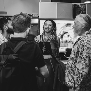 A group of diverse people are engaging in lively conversation in an indoor setting. The atmosphere is casual and friendly, with one person smiling as they speak.