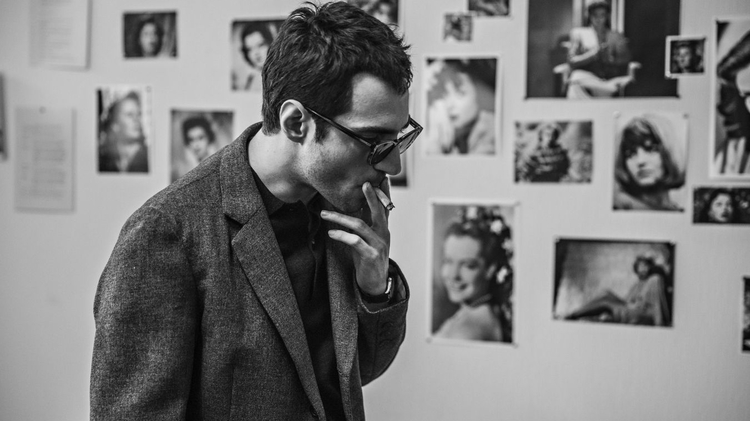 A person in a grey blazer stands pensively, surrounded by photographs pinned to a wall.