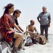 A group of four people and a dog sit on the ground in a barren landscape, surrounded by bags and belongings.