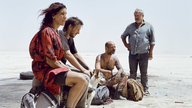 A group of four people and a dog sit on the ground in a barren landscape, surrounded by bags and belongings.