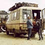Two individuals converse near a vintage van on a rocky terrain, while another person sits nearby. A second vehicle is partially visible in the background.