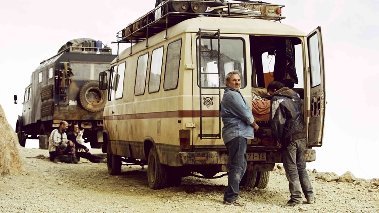 Two individuals converse near a vintage van on a rocky terrain, while another person sits nearby. A second vehicle is partially visible in the background.