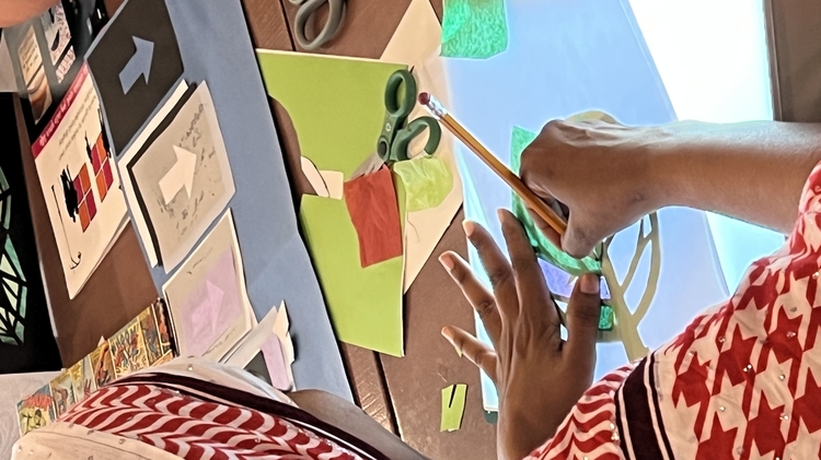 A person in a red and white patterned headscarf works on colourful paper crafts using a lightbox. Scissors and paper are scattered on the table.