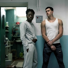 Two individuals stand in a dimly lit hallway, one in a grey sweatsuit and the other in a white tank top and dark trousers.