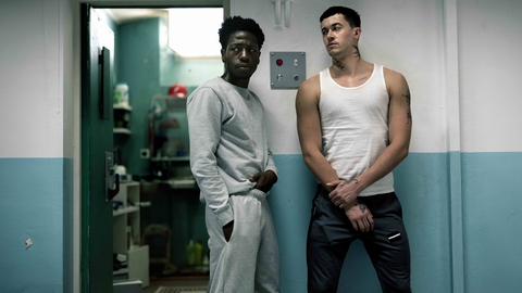 Two individuals stand in a dimly lit hallway, one in a grey sweatsuit and the other in a white tank top and dark trousers.