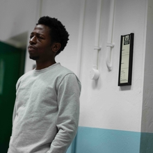 A person in a grey sweatshirt stands against a wall, beside a green door and a framed document on the wall.