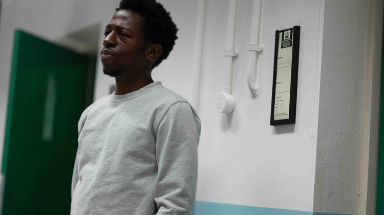 A person in a grey sweatshirt stands against a wall, beside a green door and a framed document on the wall.