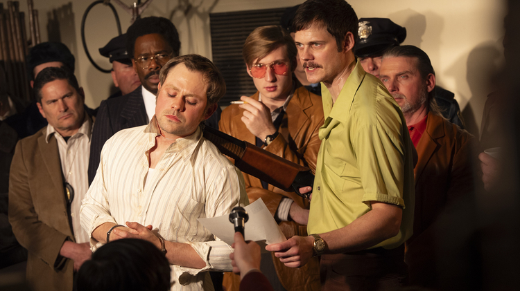 A tense scene showing a group of men in vintage attire, with one man being interviewed while others observe closely in a dimly lit room.