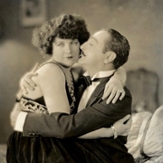 A couple in vintage attire shares a romantic embrace, sitting closely on a plush couch, radiating affection.