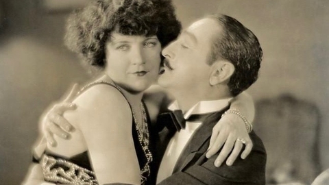 A couple in vintage attire shares a romantic embrace, sitting closely on a plush couch, radiating affection.