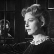 A vintage black-and-white photo of a woman in a dark, elegant dress with a lace collar, standing near a window.