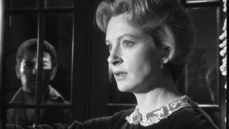 A vintage black-and-white photo of a woman in a dark, elegant dress with a lace collar, standing near a window.