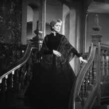 A woman in a vintage black gown and shawl poses on a wooden staircase, surrounded by ornate decor in a historic setting.