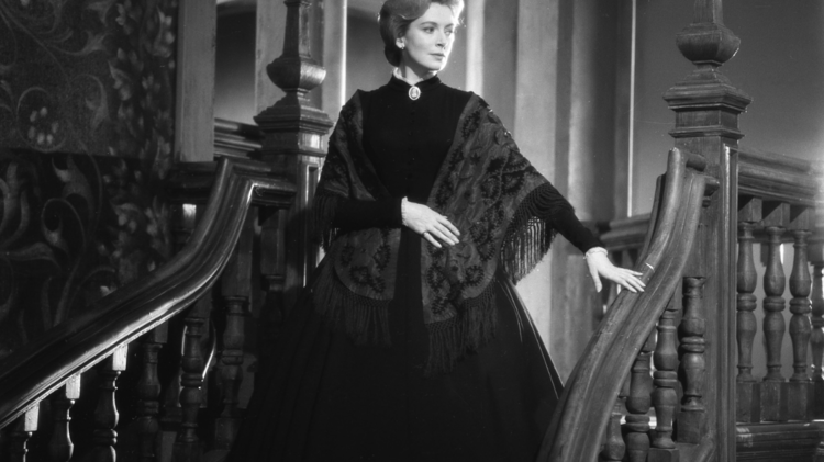 A woman in a vintage black gown and shawl poses on a wooden staircase, surrounded by ornate decor in a historic setting.