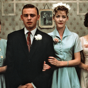 Three elegantly dressed women and a man stand together, with vintage decor in the background, exuding a classic charm.
