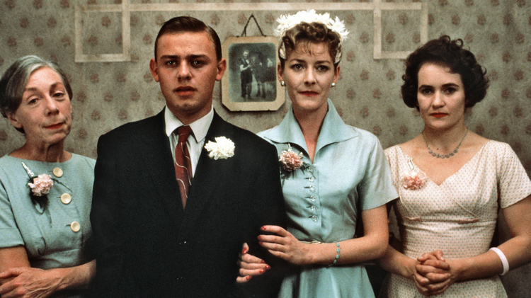 Three elegantly dressed women and a man stand together, with vintage decor in the background, exuding a classic charm.