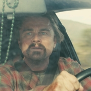A person in a plaid shirt grips the steering wheel of a car, with a blurry view through the windshield and a hanging charm in the background.