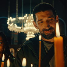 Two individuals stand in a dimly lit setting, surrounded by flickering candles, creating a warm and solemn atmosphere.