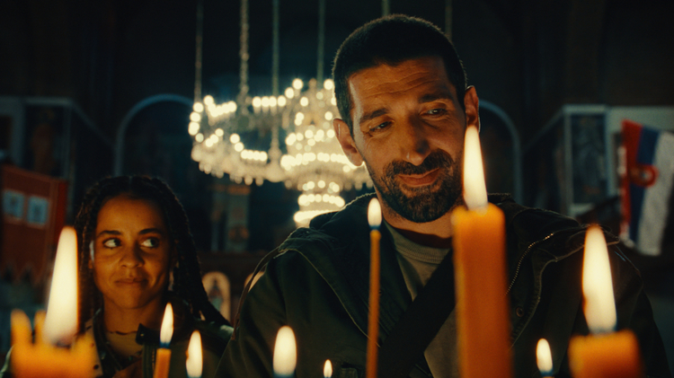 Two individuals stand in a dimly lit setting, surrounded by flickering candles, creating a warm and solemn atmosphere.