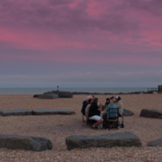 A group of people seated around a stone circle on a beach, under a vibrant pink and purple sky at dusk.