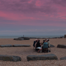 A group of people seated around a stone circle on a beach, under a vibrant pink and purple sky at dusk.