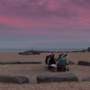 A group of people seated around a stone circle on a beach, under a vibrant pink and purple sky at dusk.